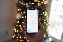 A hand holding a smartphone displaying a stock market chart with candlesticks and line graphs. In the background, there are blurred Christmas tree lights creating a festive ambiance.