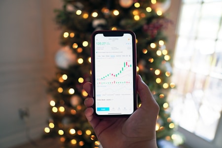 A hand holding a smartphone displaying a stock market chart with candlesticks and line graphs. In the background, there are blurred Christmas tree lights creating a festive ambiance.