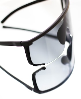 A pair of sleek, black sunglasses with tinted lenses is placed on a reflective surface. The sunglasses' modern design is emphasized by their minimalistic frame and large, rectangular lenses. The reflection creates a doubling effect, enhancing the visual impact.