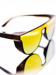 A pair of sunglasses with yellow tinted lenses and a dark brown frame. The design features perforated details on the frame corner near the temples. The reflective surface beneath them creates a mirrored effect, enhancing the visual impact.