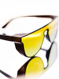 A pair of sunglasses with yellow tinted lenses and a dark brown frame. The design features perforated details on the frame corner near the temples. The reflective surface beneath them creates a mirrored effect, enhancing the visual impact.