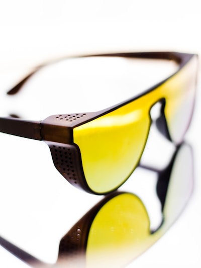 A pair of sunglasses with yellow tinted lenses and a dark brown frame. The design features perforated details on the frame corner near the temples. The reflective surface beneath them creates a mirrored effect, enhancing the visual impact.