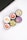 Five cosmetic containers with vivid and colorful labels depicting fruits and flowers are arranged in a cluster. The labels include images of a strawberry, peach, citrus fruits, a floral design with vanilla, and various berries. The setting is minimalistic, with a white surface and part of an electronic device visible in the corner.