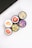 Five cosmetic containers with vivid and colorful labels depicting fruits and flowers are arranged in a cluster. The labels include images of a strawberry, peach, citrus fruits, a floral design with vanilla, and various berries. The setting is minimalistic, with a white surface and part of an electronic device visible in the corner.
