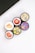 Five cosmetic containers with vivid and colorful labels depicting fruits and flowers are arranged in a cluster. The labels include images of a strawberry, peach, citrus fruits, a floral design with vanilla, and various berries. The setting is minimalistic, with a white surface and part of an electronic device visible in the corner.