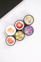 Five cosmetic containers with vivid and colorful labels depicting fruits and flowers are arranged in a cluster. The labels include images of a strawberry, peach, citrus fruits, a floral design with vanilla, and various berries. The setting is minimalistic, with a white surface and part of an electronic device visible in the corner.