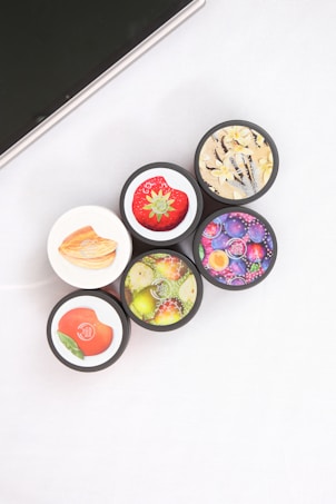 Five cosmetic containers with vivid and colorful labels depicting fruits and flowers are arranged in a cluster. The labels include images of a strawberry, peach, citrus fruits, a floral design with vanilla, and various berries. The setting is minimalistic, with a white surface and part of an electronic device visible in the corner.
