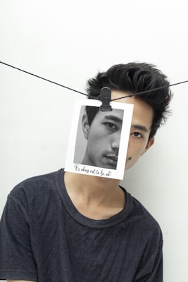 A person wearing a dark shirt appears to be partially covered by a monochrome photograph clipped to a line, obscuring part of their face. The photo has text saying 'It's okay not to be ok!' The background is plain white, and the line is strung horizontally across the image.