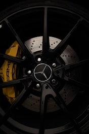A close-up view of a car wheel, specifically featuring a black alloy rim with a silver emblem at the center. The rim is fitted with a high-performance yellow brake caliper adorned with branding. The brake disc has visible drilled holes, emphasizing its sporty design.
