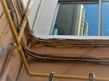 A view of an exterior wall with multiple electrical cables and pipes running along it. The wall is light brown, and the cables are arranged in a slightly chaotic manner. A yellow pipe stands out prominently. Above the wall, a window reflects a portion of a building, showing a bright blue sky and the facade of a high-rise structure.