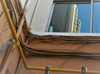 A view of an exterior wall with multiple electrical cables and pipes running along it. The wall is light brown, and the cables are arranged in a slightly chaotic manner. A yellow pipe stands out prominently. Above the wall, a window reflects a portion of a building, showing a bright blue sky and the facade of a high-rise structure.