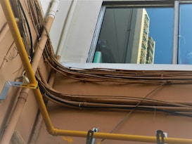 A view of an exterior wall with multiple electrical cables and pipes running along it. The wall is light brown, and the cables are arranged in a slightly chaotic manner. A yellow pipe stands out prominently. Above the wall, a window reflects a portion of a building, showing a bright blue sky and the facade of a high-rise structure.