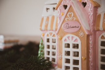 A ceramic decorative house resembling a patisserie, featuring pastel pink and white colors with heart and floral motifs. The structure has intricate windows and small details that give it a quaint, charming appearance. Spruce branches are positioned at the base, enhancing the festive aesthetic.