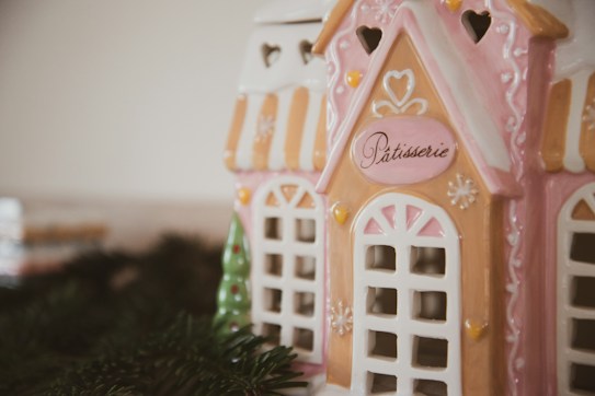 A ceramic decorative house resembling a patisserie, featuring pastel pink and white colors with heart and floral motifs. The structure has intricate windows and small details that give it a quaint, charming appearance. Spruce branches are positioned at the base, enhancing the festive aesthetic.