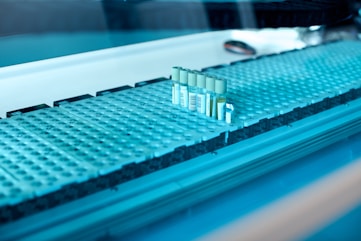 A laboratory setting featuring a conveyor belt with several test tubes placed upright in holders. The tubes are arranged in a line and have labels with barcodes. The lighting creates a cool blue tone across the surface, suggesting a sterile and clean environment.