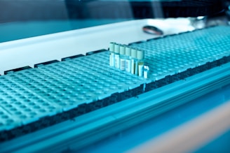 A laboratory setting featuring a conveyor belt with several test tubes placed upright in holders. The tubes are arranged in a line and have labels with barcodes. The lighting creates a cool blue tone across the surface, suggesting a sterile and clean environment.