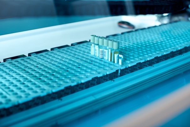 A laboratory setting featuring a conveyor belt with several test tubes placed upright in holders. The tubes are arranged in a line and have labels with barcodes. The lighting creates a cool blue tone across the surface, suggesting a sterile and clean environment.