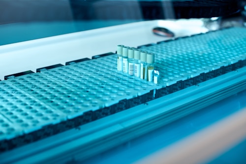 A laboratory setting featuring a conveyor belt with several test tubes placed upright in holders. The tubes are arranged in a line and have labels with barcodes. The lighting creates a cool blue tone across the surface, suggesting a sterile and clean environment.