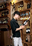 A person is examining a bottle of wine in a well-stocked wine cellar. Wooden shelves display various bottles of liquor, and warm lighting creates a cozy atmosphere.
