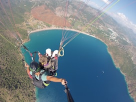Aerial view of two people paragliding over a vibrant blue body of water, with a scenic landscape featuring green forests, mountains, and a sandy shoreline.