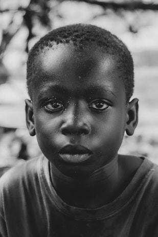 A black and white portrait of a young child with a neutral expression. The background is blurred, drawing focus to the child's detailed facial features and expressive eyes.