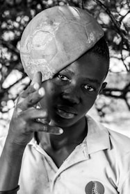 A young person is playfully balancing a worn soccer ball on their head. Their gaze is direct and intense, while their hand gestures in a playful manner. The image is black and white, highlighting the texture of the ball and the person's clothing, which features a logo.