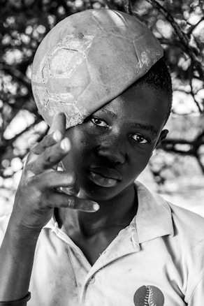 A young person is playfully balancing a worn soccer ball on their head. Their gaze is direct and intense, while their hand gestures in a playful manner. The image is black and white, highlighting the texture of the ball and the person's clothing, which features a logo.