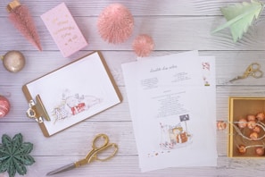 A flat lay composition featuring a clipboard with a Christmas-themed illustration, a recipe sheet, pink bottle brush trees, gold scissors, a pink greeting card, a green paper tree, a green geometric leaf decoration, a gold box with string lights, and various festive ornaments all arranged on a light wooden surface.