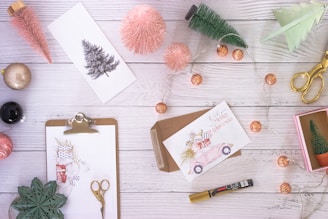 An arrangement of festive items on a white wooden surface includes Christmas cards, decorative trees, a pink glittery ornament, scissors, a marker, and a string of fairy lights. A clipboard holds a festive illustration with holiday motifs, and a pair of gold scissors lies nearby. The scene evokes cozy, creative holiday preparation.