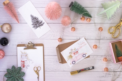 An arrangement of festive items on a white wooden surface includes Christmas cards, decorative trees, a pink glittery ornament, scissors, a marker, and a string of fairy lights. A clipboard holds a festive illustration with holiday motifs, and a pair of gold scissors lies nearby. The scene evokes cozy, creative holiday preparation.