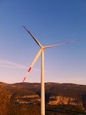 A wind turbine with three blades stands tall against a clear blue sky. The turbine's blades have red markings and it is situated in a hilly landscape with brown and green foliage, likely during sunset, giving the scene a warm glow. The background consists of mountains with patches of vegetation.