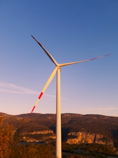 A wind turbine with three blades stands tall against a clear blue sky. The turbine's blades have red markings and it is situated in a hilly landscape with brown and green foliage, likely during sunset, giving the scene a warm glow. The background consists of mountains with patches of vegetation.