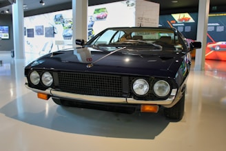 A vintage sports car with a sleek, dark exterior is displayed in a modern showroom. It has two round headlights, a distinctive grille, and chrome accents. The surroundings include informational displays and images related to automotive history.