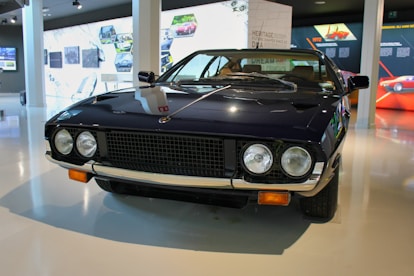 A vintage sports car with a sleek, dark exterior is displayed in a modern showroom. It has two round headlights, a distinctive grille, and chrome accents. The surroundings include informational displays and images related to automotive history.