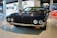 A vintage sports car with a sleek, dark exterior is displayed in a modern showroom. It has two round headlights, a distinctive grille, and chrome accents. The surroundings include informational displays and images related to automotive history.
