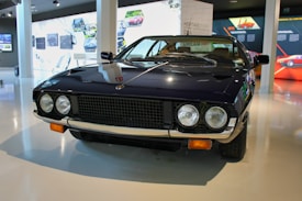 A vintage sports car with a sleek, dark exterior is displayed in a modern showroom. It has two round headlights, a distinctive grille, and chrome accents. The surroundings include informational displays and images related to automotive history.