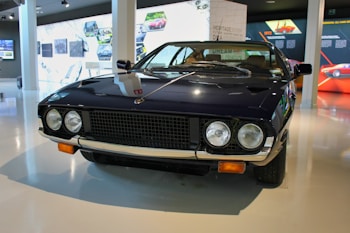 A vintage sports car with a sleek, dark exterior is displayed in a modern showroom. It has two round headlights, a distinctive grille, and chrome accents. The surroundings include informational displays and images related to automotive history.
