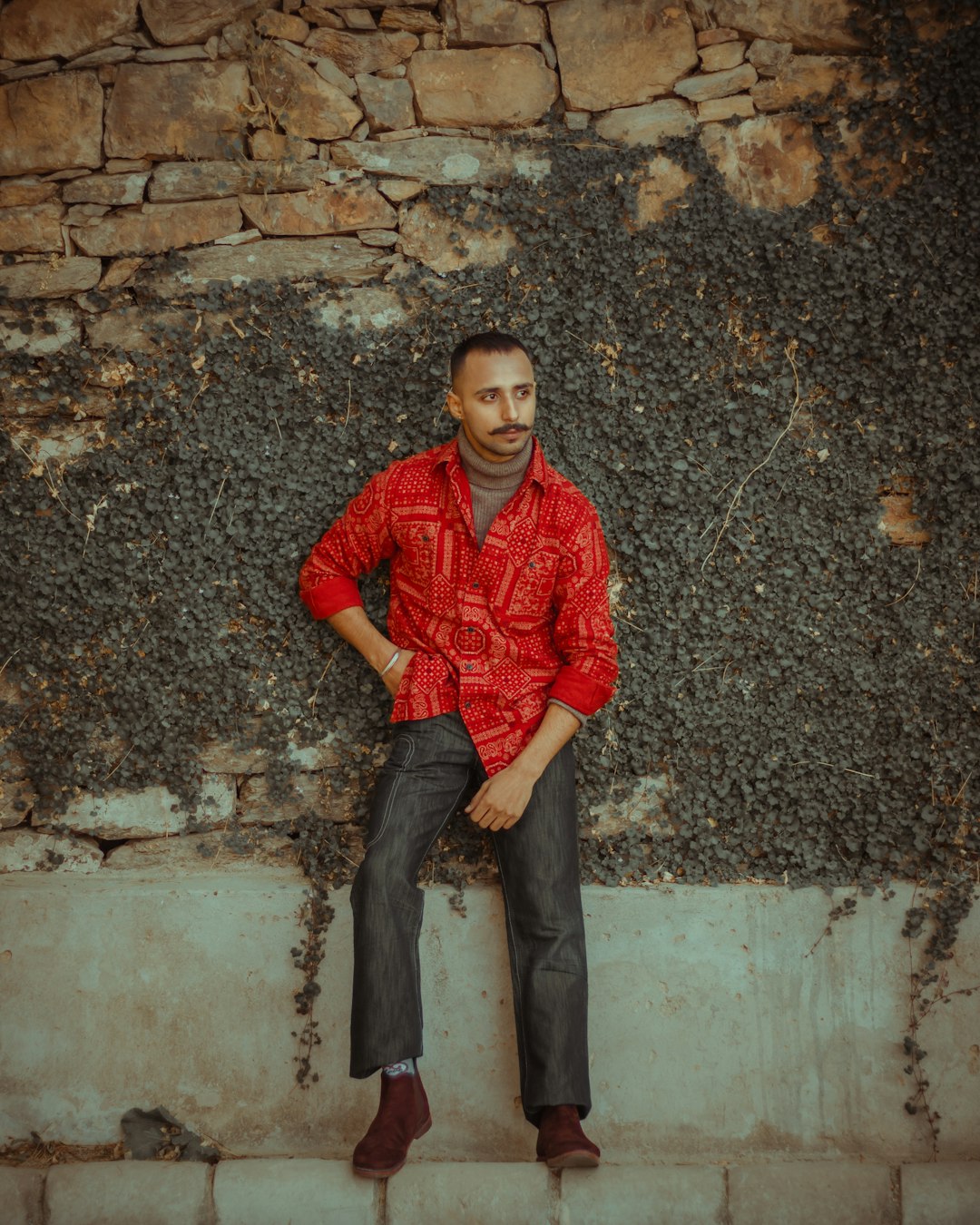 man in red and black dress shirt and black denim jeans sitting on concrete wall