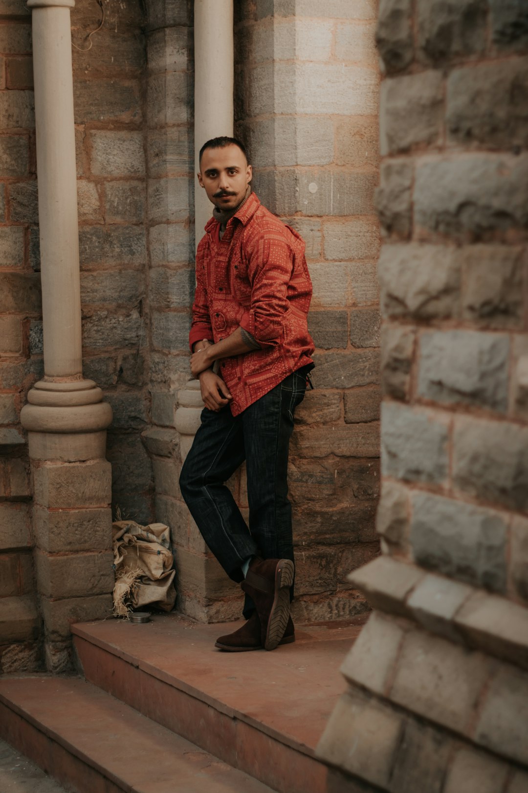 man in red jacket and black denim jeans standing beside brown concrete pillar
