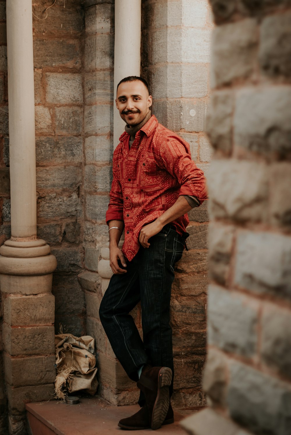 man in red dress shirt and black denim jeans sitting on concrete column