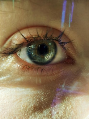 A close-up of a human eye with greenish-blue iris and visible eyelashes. The skin texture around the eye is prominent, and there are subtle reflections and linear light patterns across the eye and eyelid.