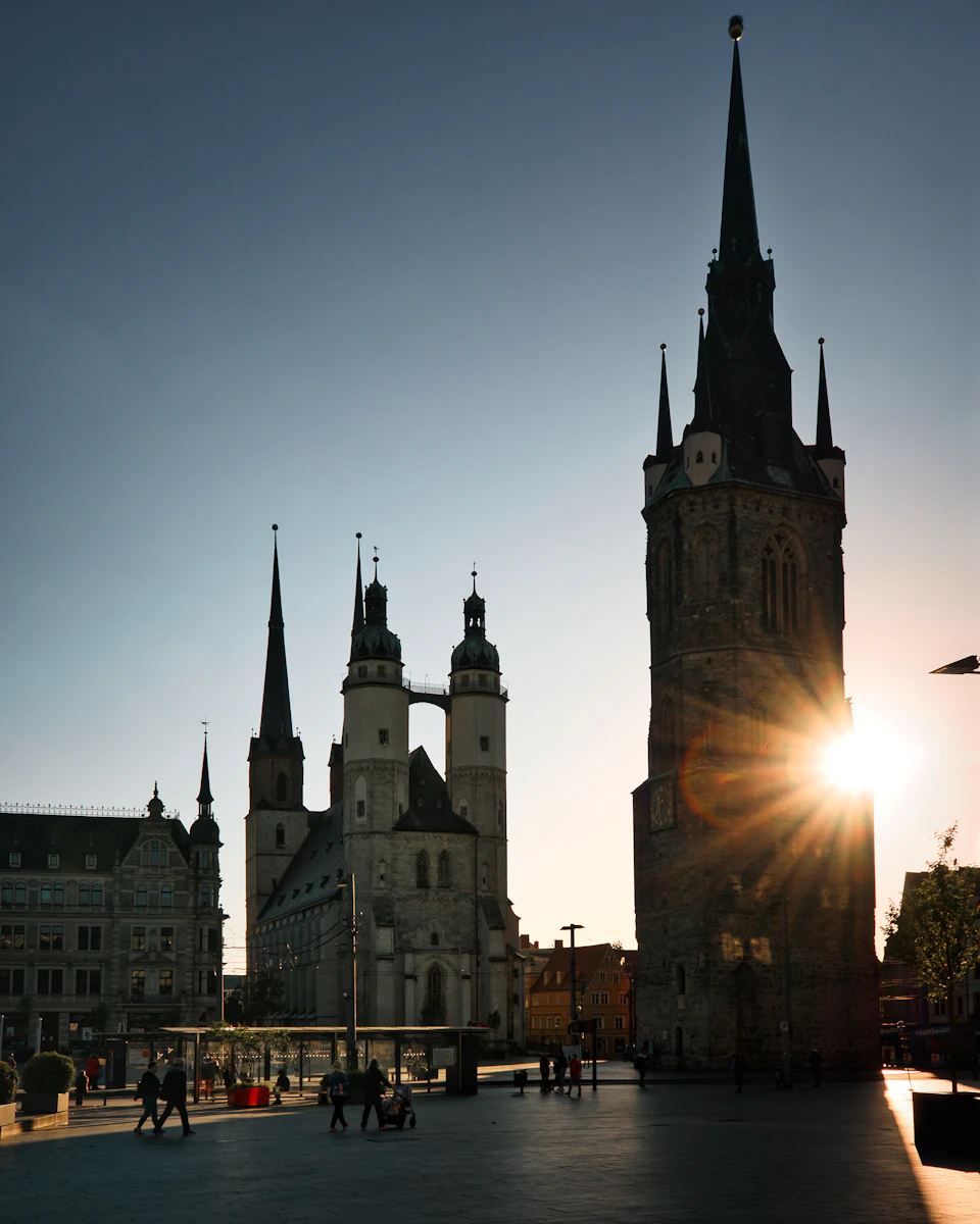 The image depicts a serene cityscape at sunset, with the sun's rays casting a warm glow over the scene. Dominating the center is a tall tower, its pointed spire reaching towards the sky. The tower is part of a larger building complex that includes several other structures, including a church and a castle-like edifice.

The city square below is bustling with activity as people walk around, some strolling leisurely while others are engaged in conversation or observing their surroundings. A few individuals can be seen sitting on benches scattered throughout the area, enjoying the tranquil atmosphere of the evening. The sky above is painted in shades of blue and orange, providing a beautiful backdrop to this picturesque scene.