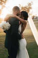 A couple embraces closely in a romantic setting, with the woman wearing a white dress and veil and holding a bouquet of white flowers. The man wears a dark suit, and they stand on a grassy area with soft, warm sunlight illuminating the scene.