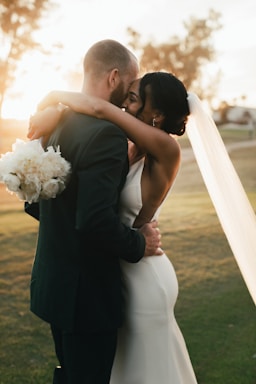 A couple embraces closely in a romantic setting, with the woman wearing a white dress and veil and holding a bouquet of white flowers. The man wears a dark suit, and they stand on a grassy area with soft, warm sunlight illuminating the scene.