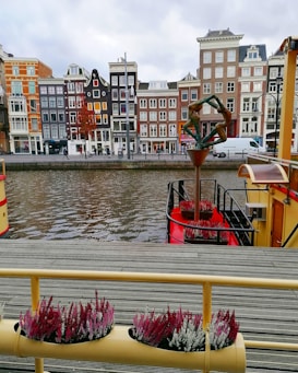 Colorful canal-side buildings with classic Dutch architecture stand along a waterway. A sculpture of two figures in a geometric pose is prominently displayed on a boat dock. The atmosphere is enhanced by flowers and a view across the water, blending a mix of natural and urban elements.