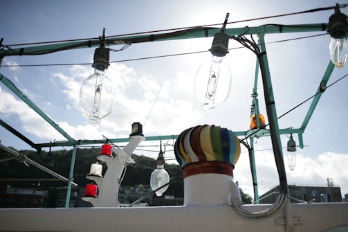 A series of large glass light fixtures are mounted on a metal frame against a bright sky. Red, white, and yellow light covers are visible on a nearby structure, with a prominent multi-colored circular element at the forefront. There is a background of distant hills and buildings partially obscured.