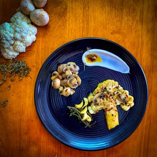 A black plate on a wooden surface presents a gourmet dish featuring a large cauliflower steak garnished with mushrooms, herbs, and red pepper flakes. Beside it, sliced mushrooms are arranged neatly. A dollop of sauce with a touch of oil is elegantly swirled on the plate. In the background, raw cauliflower, whole mushrooms, and sprigs of thyme are artfully placed.