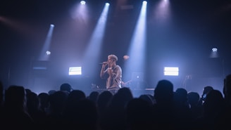 A singer performs on stage under dramatic lighting, with spotlights illuminating the area. The audience is visible in the foreground, watching the performance with interest. Equipment, such as a laptop, can be seen on a table behind the performer.