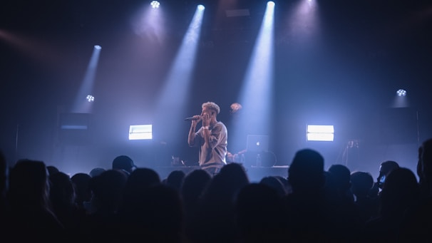 A singer performs on stage under dramatic lighting, with spotlights illuminating the area. The audience is visible in the foreground, watching the performance with interest. Equipment, such as a laptop, can be seen on a table behind the performer.