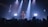 A singer performs on stage under dramatic lighting, with spotlights illuminating the area. The audience is visible in the foreground, watching the performance with interest. Equipment, such as a laptop, can be seen on a table behind the performer.
